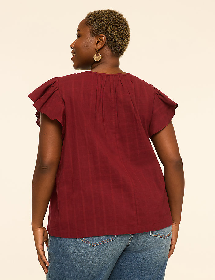A woman in a maroon flutter-sleeve top and blue jeans stands against a plain, light-colored background.