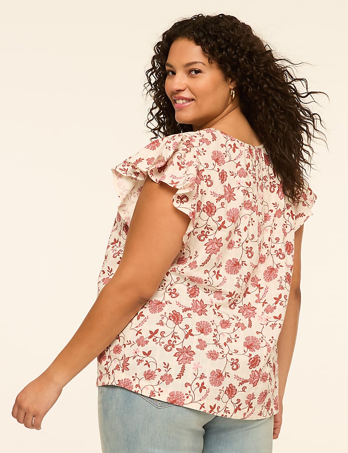 A woman with curly dark hair smiles over her shoulder, wearing a floral print blouse and light blue jeans against a plain cream background.