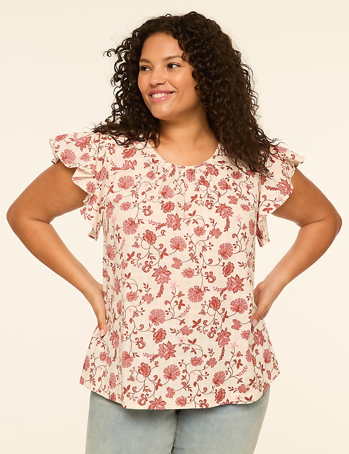 A smiling woman with curly dark hair poses with her hands on her hips, wearing a cream-colored floral print top with ruffled sleeves, against a plain cream background.