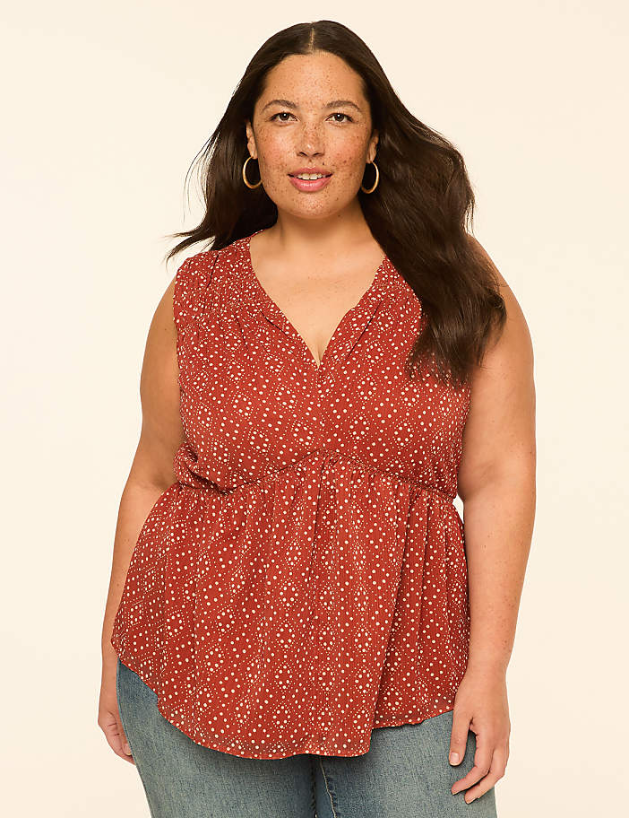 A woman with dark hair and freckles, wearing a rust-colored patterned top and blue jeans, smiles against a plain, light-colored background.