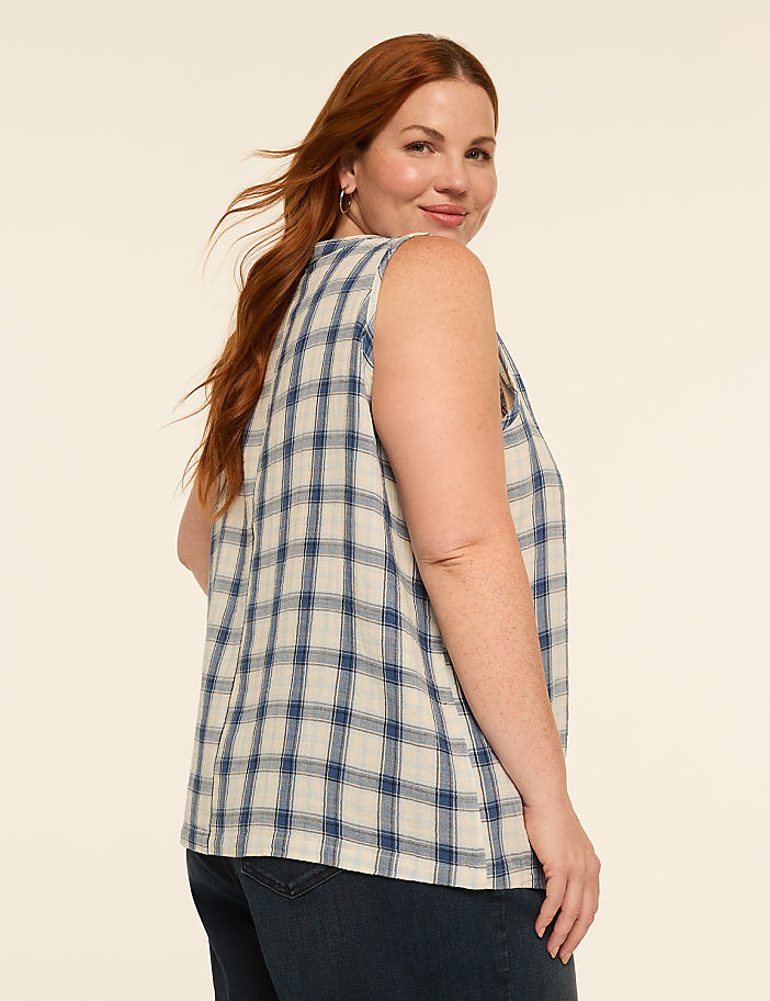 A woman with red hair wearing a plaid sleeveless top and jeans poses against a plain, light-colored background.