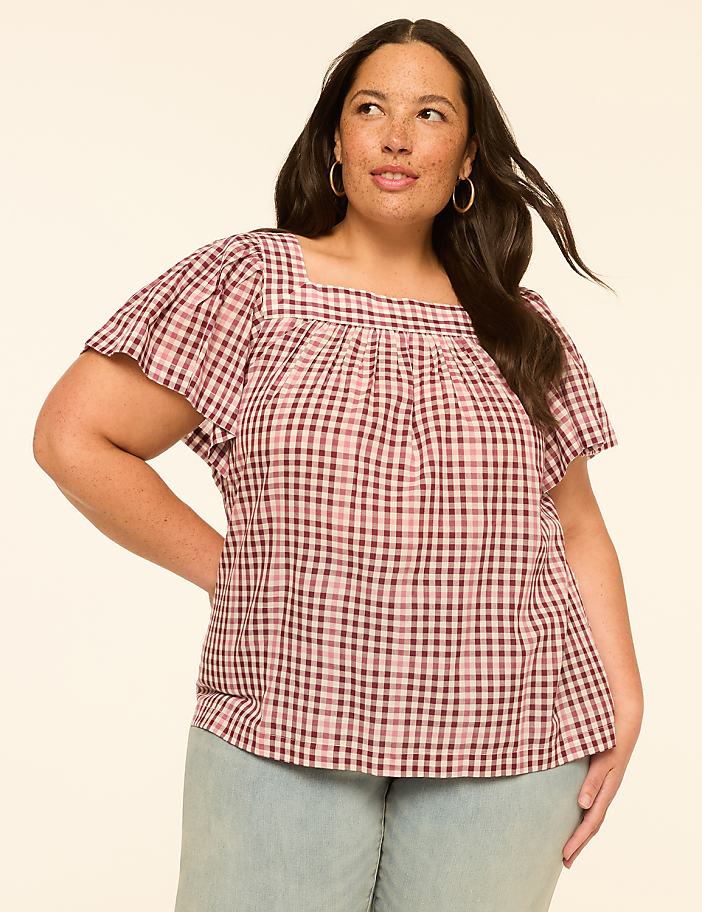 A woman with dark hair and freckles poses in a pink gingham top and light blue pants against a plain, light-colored background.