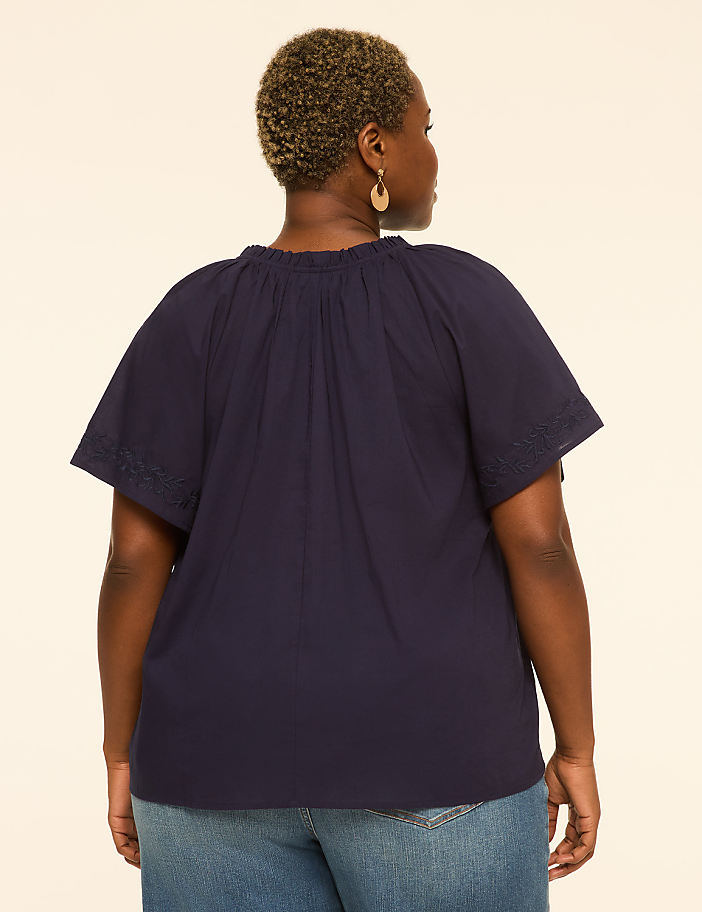 A woman in a navy blue top and jeans stands against a plain, light-colored background.