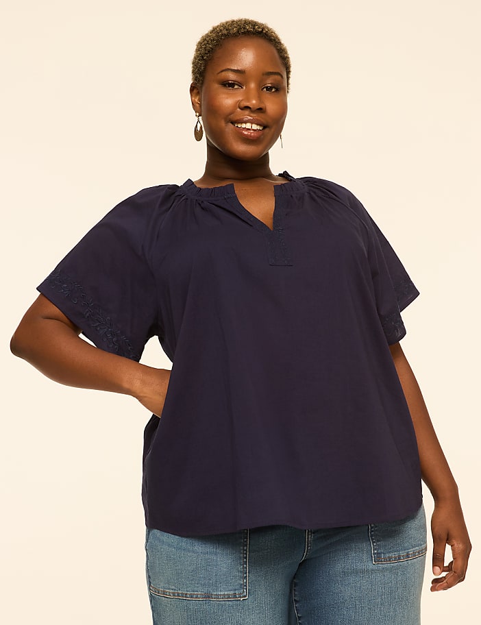 A smiling woman in a navy blue top and jeans stands against a plain, light-colored background.