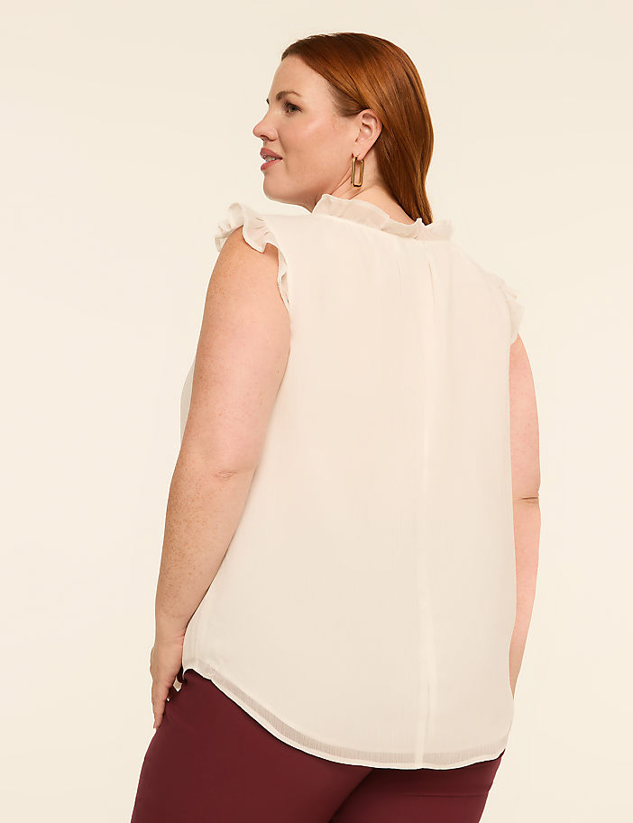 A woman with red hair, wearing a cream-colored ruffled blouse and maroon pants, poses against a plain, light-colored background.