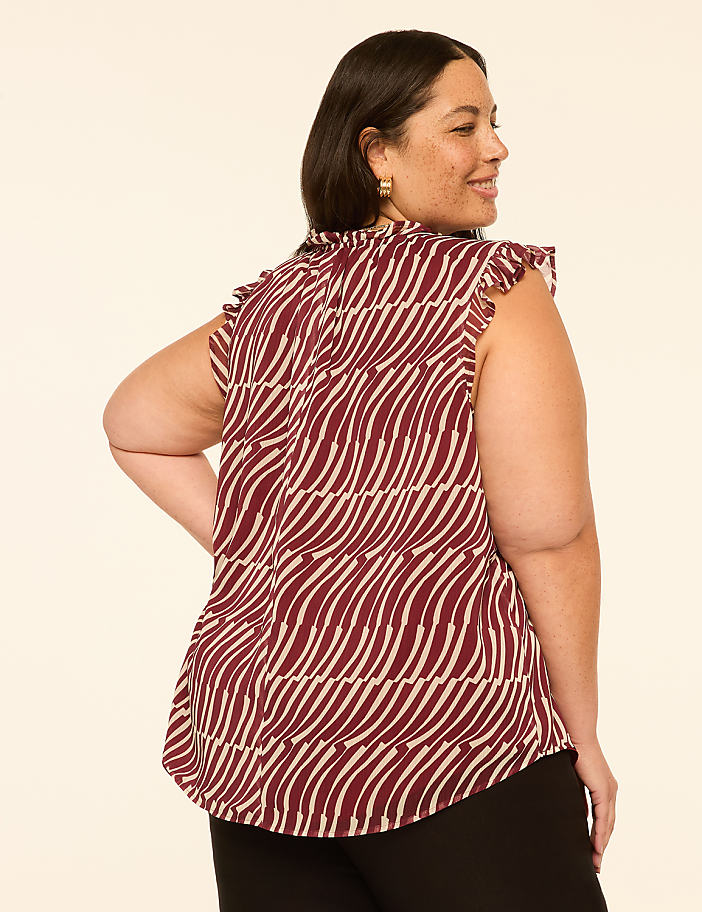 A woman in a patterned maroon top and black pants stands against a plain cream background.
