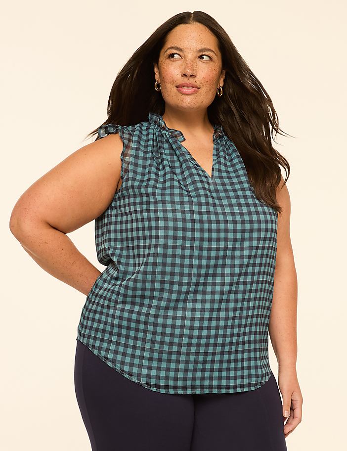 A woman with dark hair and freckles poses in a teal and navy gingham top with ruffled sleeves, set against a plain, light beige background.