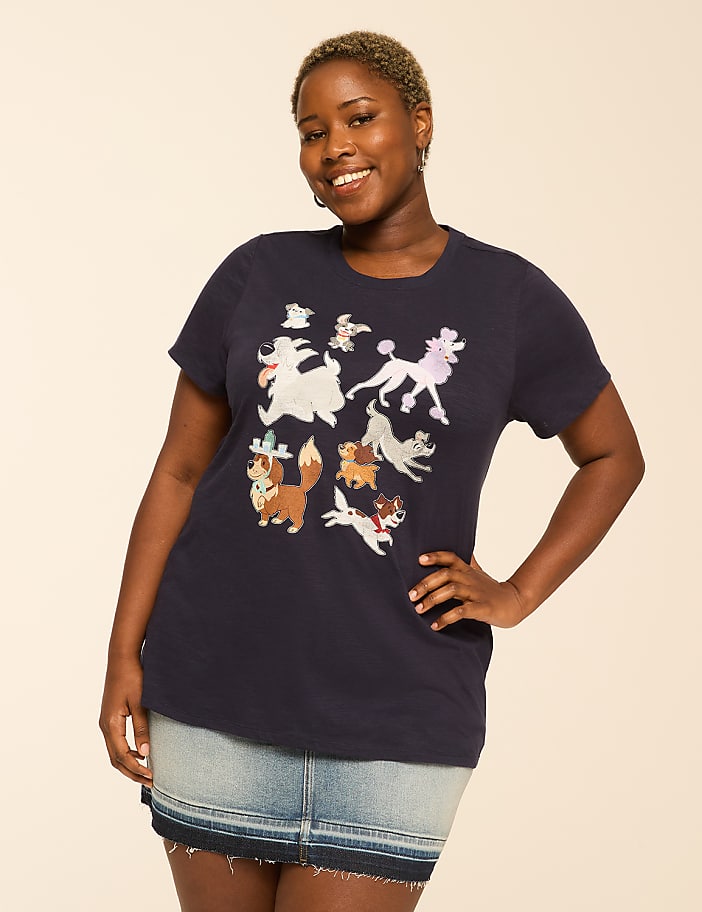 A smiling woman in a dark blue t-shirt with cartoon dogs and a denim skirt stands against a plain, light-colored background.