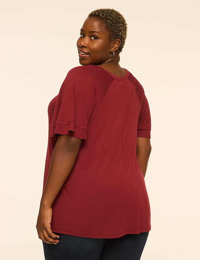 A woman in a maroon shirt and dark jeans stands against a plain, light-colored background.