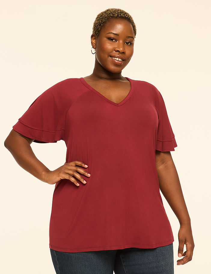 A smiling woman with short, dark hair and a maroon v-neck shirt stands with her hand on her hip against a plain, light-colored background.