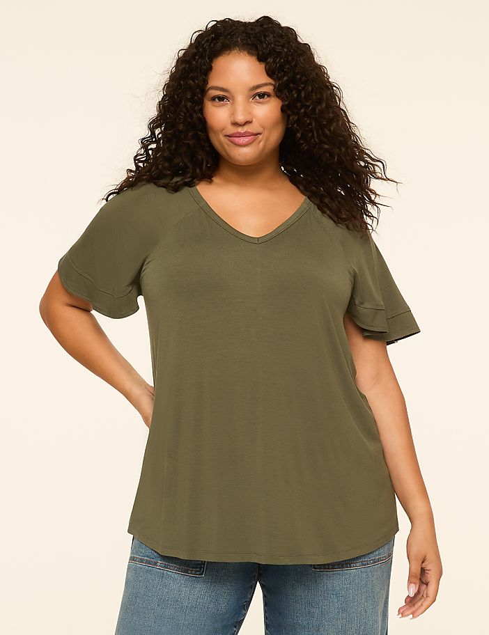 A woman with curly dark hair and a v-neck olive green t-shirt stands against a plain cream background.