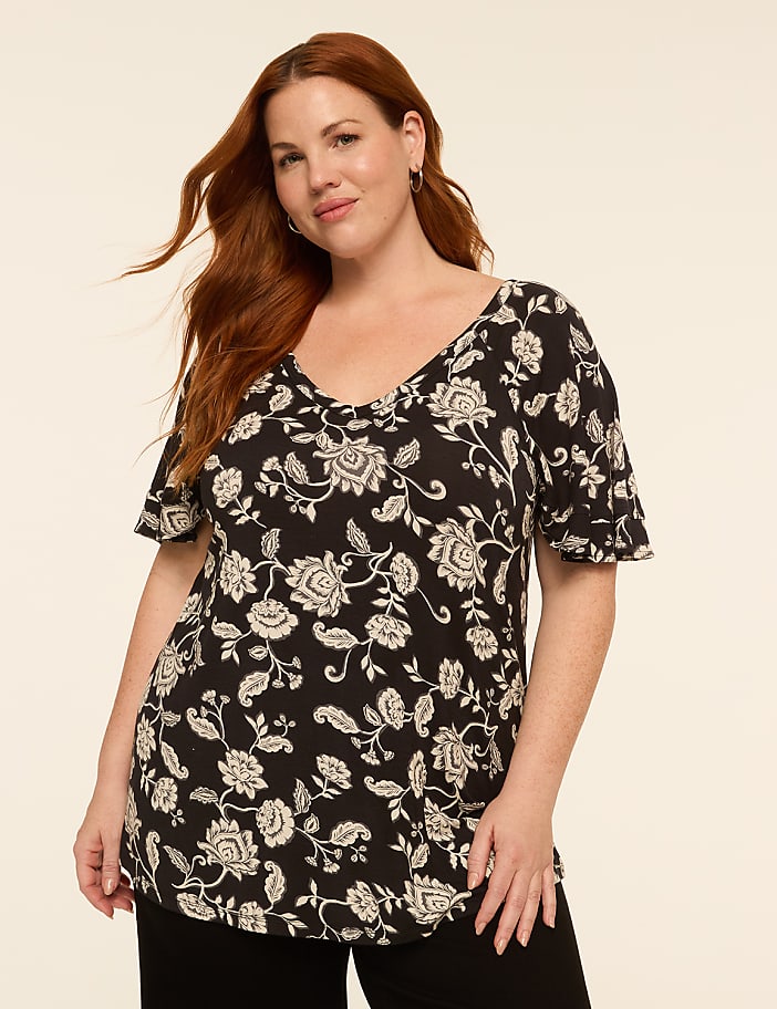A woman with auburn hair and a floral print top stands against a plain, light-colored background.