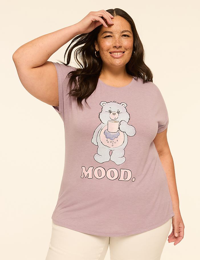 A smiling woman in a purple t-shirt with a cartoon bear and the word "MOOD" stands against a plain, light-colored background.