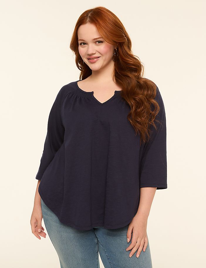 A smiling woman with long, wavy red hair and a navy blue top stands against a plain, light-colored background.