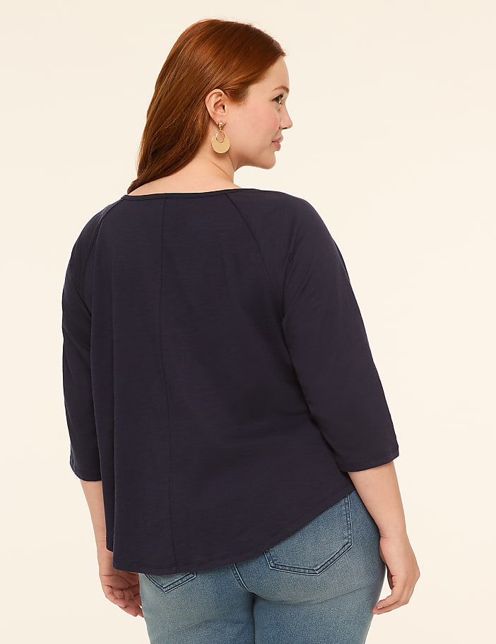 A woman with red hair, wearing a navy blue shirt and jeans, stands against a plain, light-colored background.