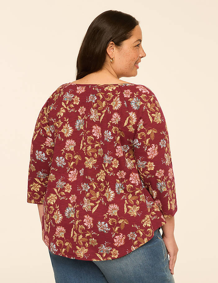 A woman in a floral-patterned maroon top and blue jeans stands against a plain, light-colored background.