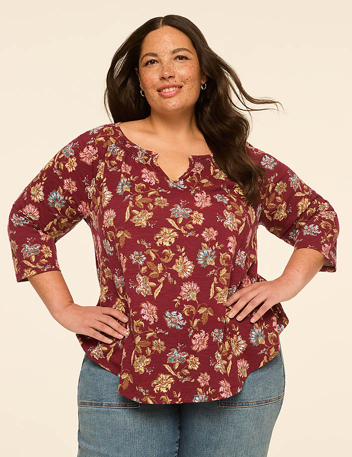 A woman with dark hair and freckles smiles while wearing a floral-patterned maroon top and blue jeans against a plain beige background.