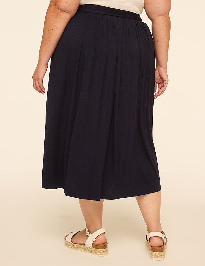 A person wearing a dark blue pleated skirt and white sandals stands against a plain beige background.