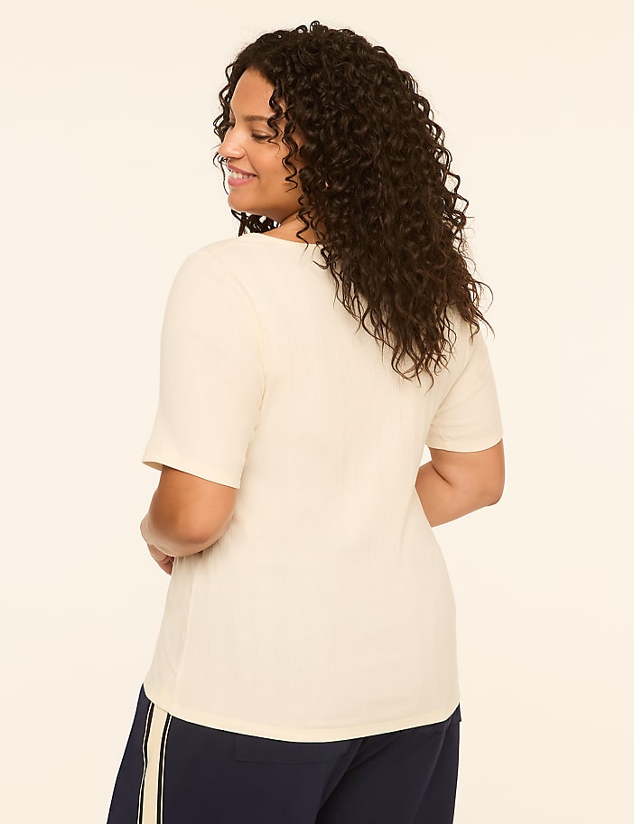 A woman in a cream-colored t-shirt and navy pants stands against a plain, light-colored background.