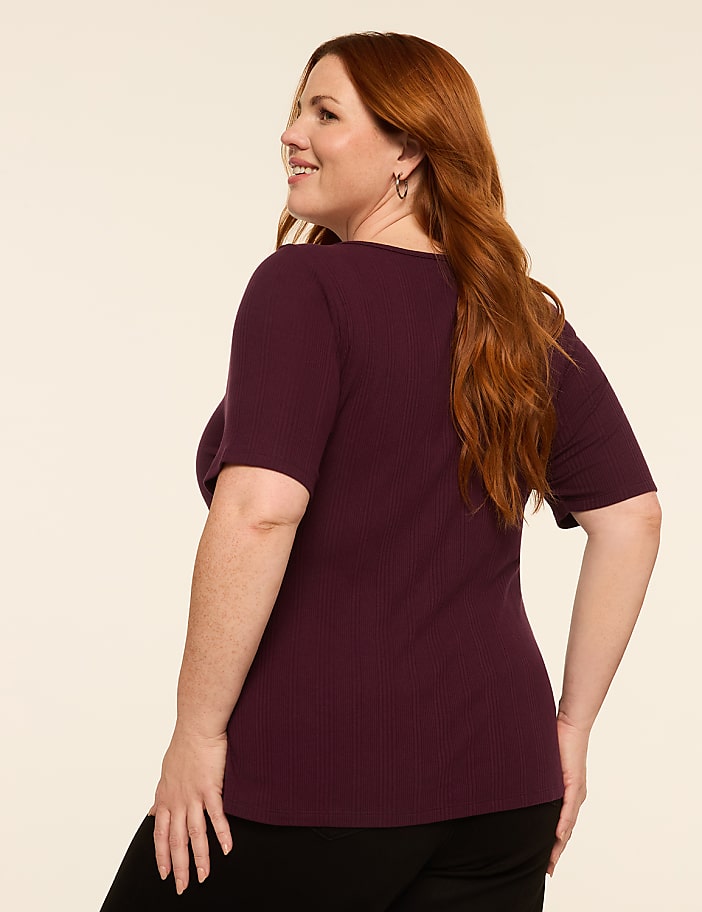 A woman with long, wavy red hair, wearing a burgundy ribbed t-shirt and black pants, smiles as she poses against a plain, light-colored background.