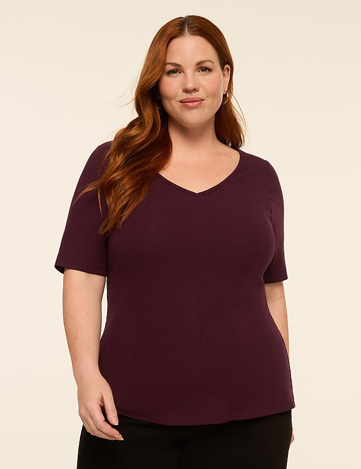 A woman with auburn hair and a v-neck shirt stands against a plain, light-colored background.