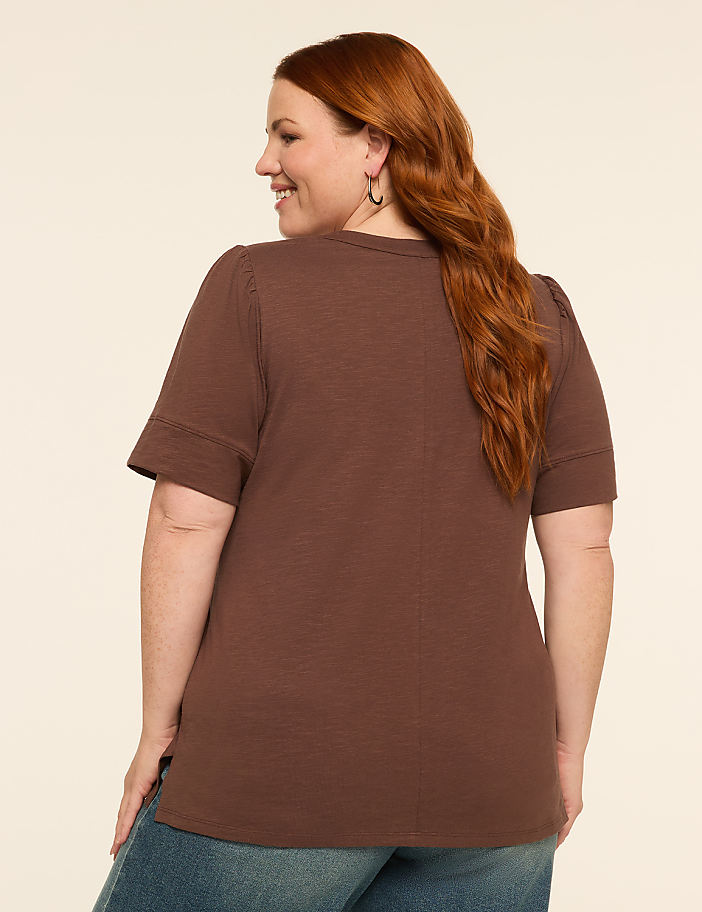 A woman with auburn hair, wearing a brown t-shirt and blue jeans, smiles as she stands against a plain, light-colored background.