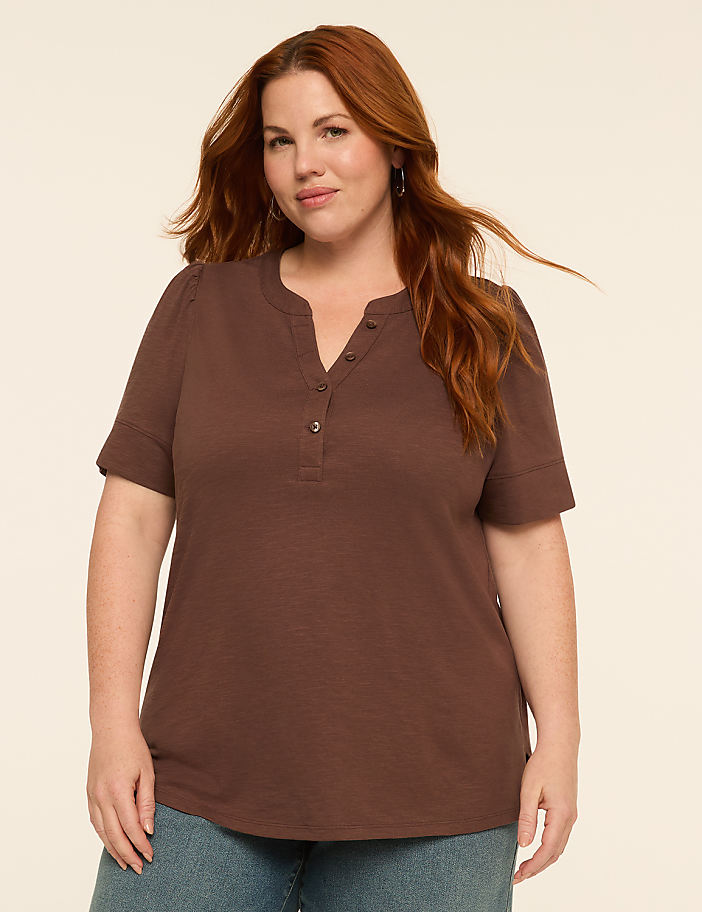 A woman with auburn hair and a brown shirt stands against a plain, light-colored background.