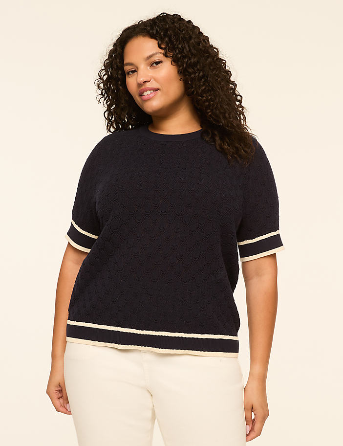 A plus-size woman with curly dark hair poses in a navy blue short-sleeved sweater with cream trim against a plain cream background.