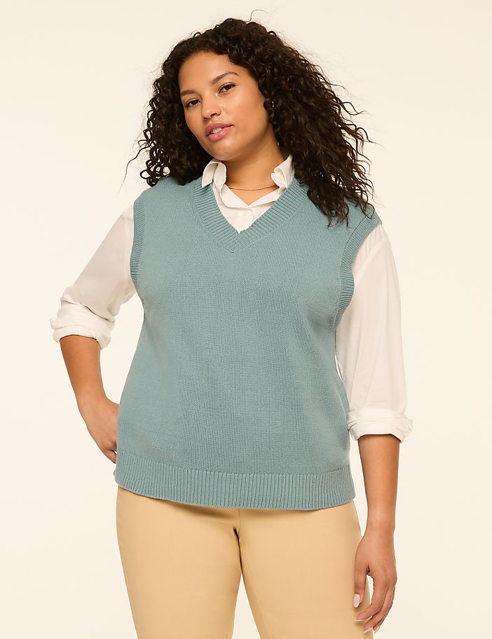 A woman in a teal v-neck sweater vest over a white collared shirt and tan pants stands against a plain cream background.