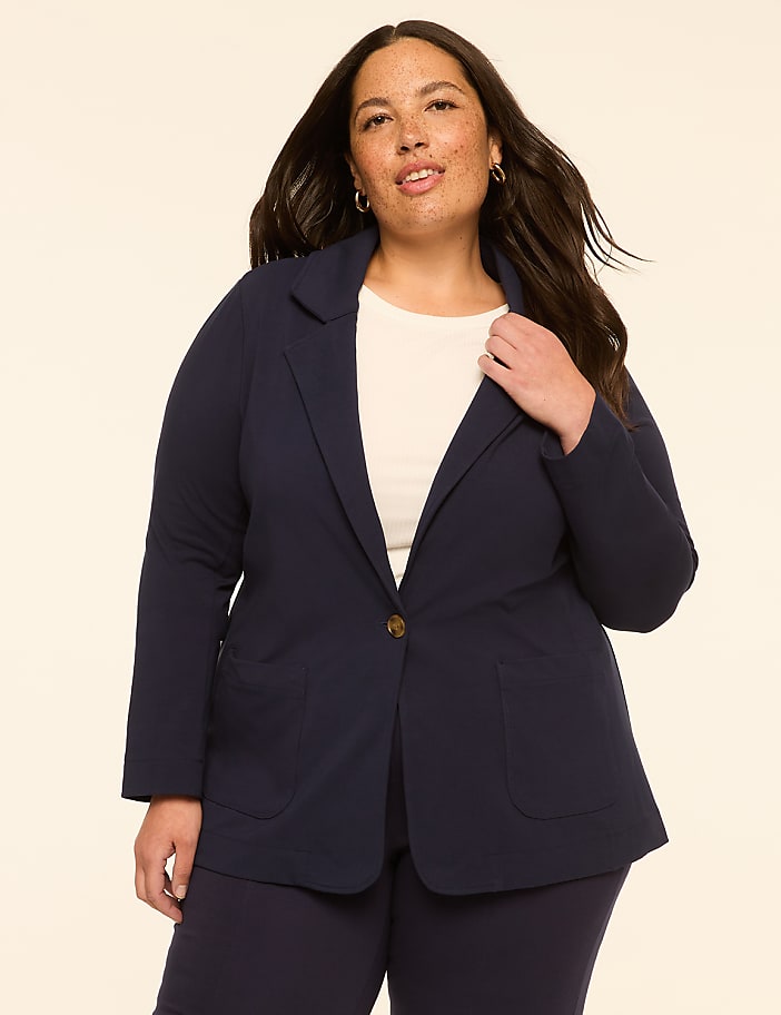 A woman in a navy blazer stands against a plain, light-colored background.