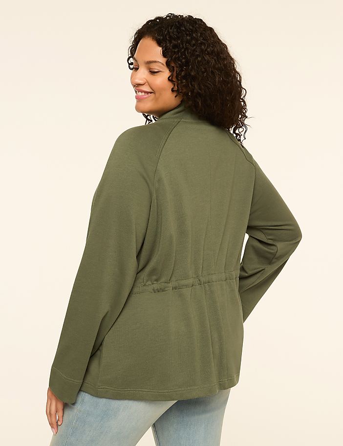 A woman in a green jacket and jeans stands against a plain, light-colored background.
