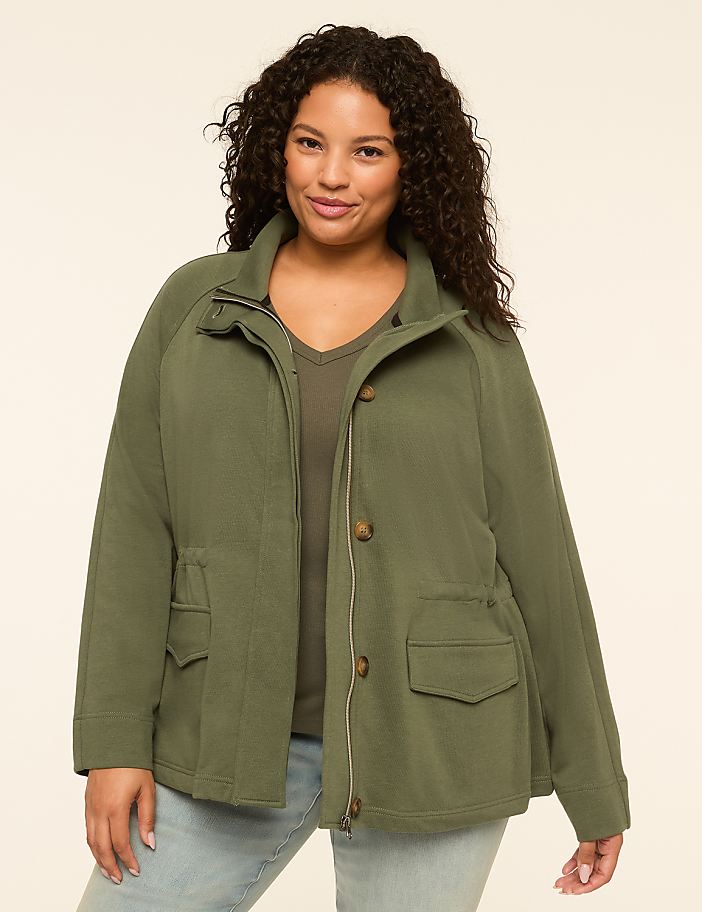 A smiling woman with curly dark hair stands in the foreground wearing a green jacket and light blue jeans, against a plain, light beige background.