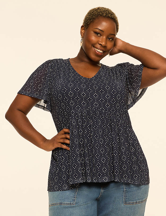 A smiling woman with short, blonde hair and a patterned blue top poses with her hand on her hip against a plain, light-colored background.