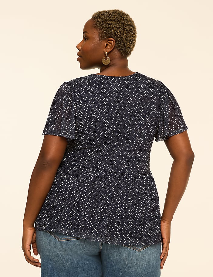 A woman in a patterned navy blouse and jeans stands against a plain, light-colored background.