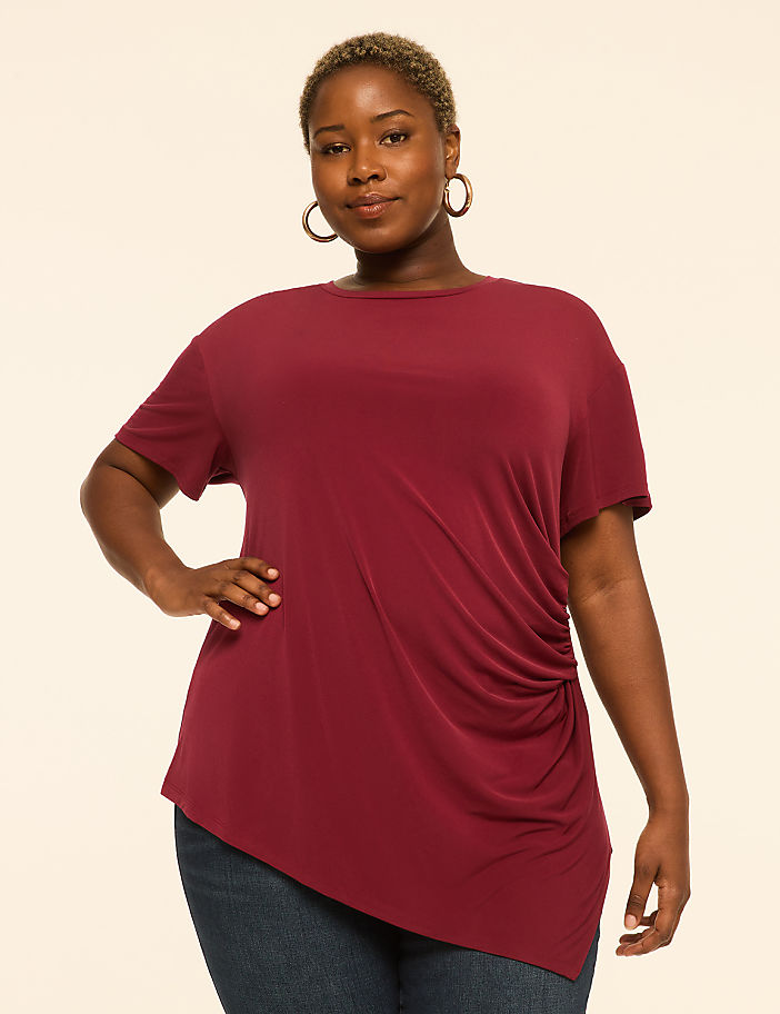A woman in a maroon tunic and blue jeans stands against a plain beige background.