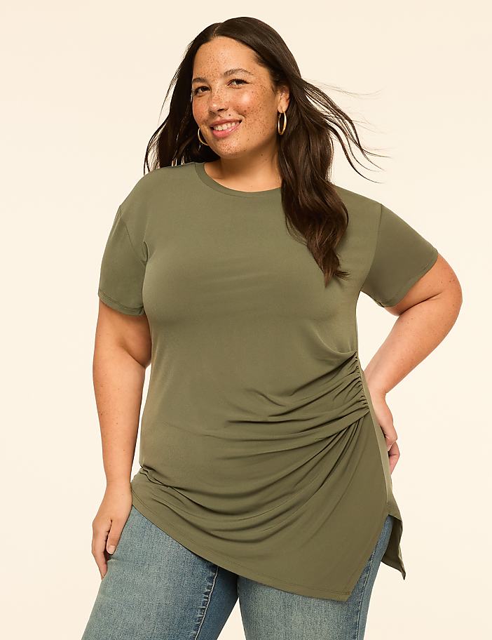 A smiling woman in a ruched olive green top and jeans stands against a plain, light-colored background.