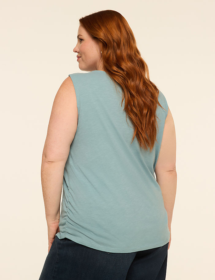 A woman with red hair, wearing a teal tank top and dark jeans, stands against a plain, light-colored background.