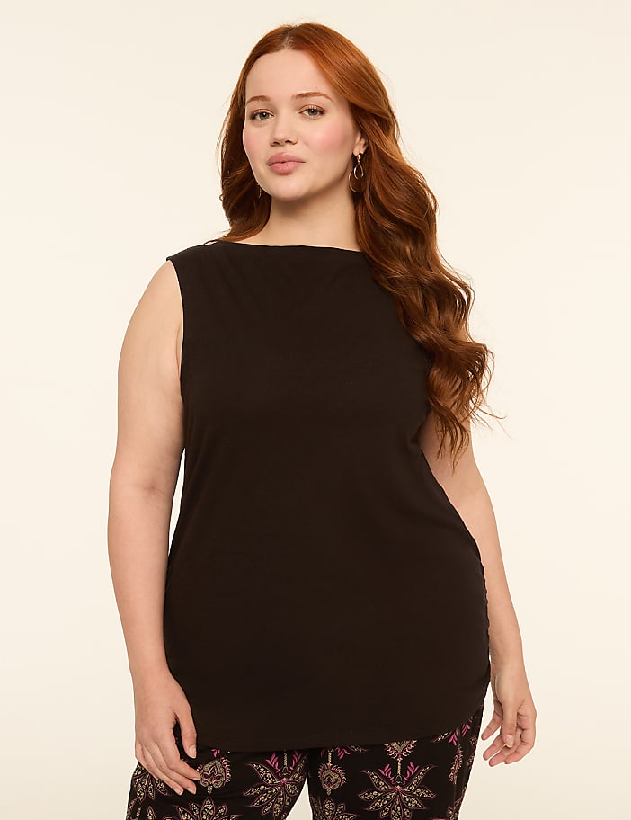 A woman with long, wavy red hair and a black sleeveless top stands against a plain, light-colored background, wearing patterned pants.