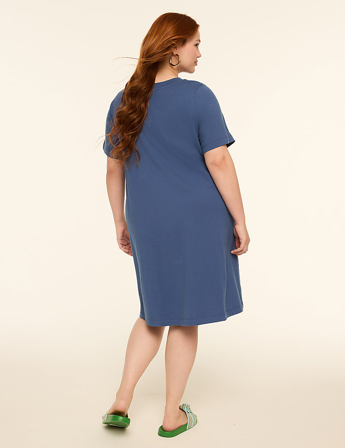 A woman with long red hair, wearing a blue t-shirt dress and green slides, stands against a plain, light-colored background.