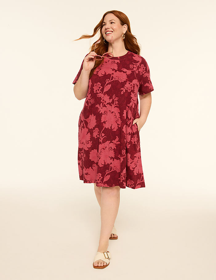 A smiling woman in a floral dress walks forward against a plain, light-colored background.