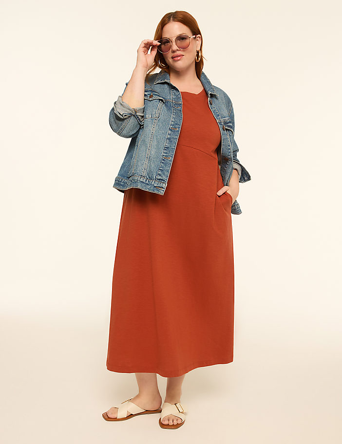 A woman in a rust-colored dress and denim jacket stands against a plain, light-colored background.
