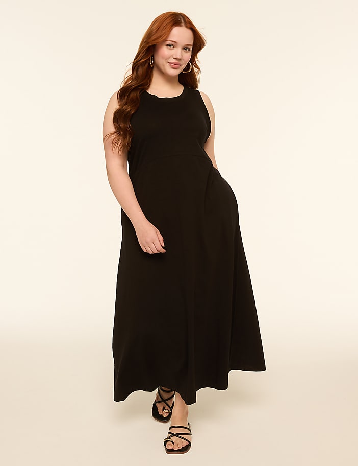 A woman with long, wavy red hair stands in a simple black maxi dress and black sandals against a plain, light-colored background.