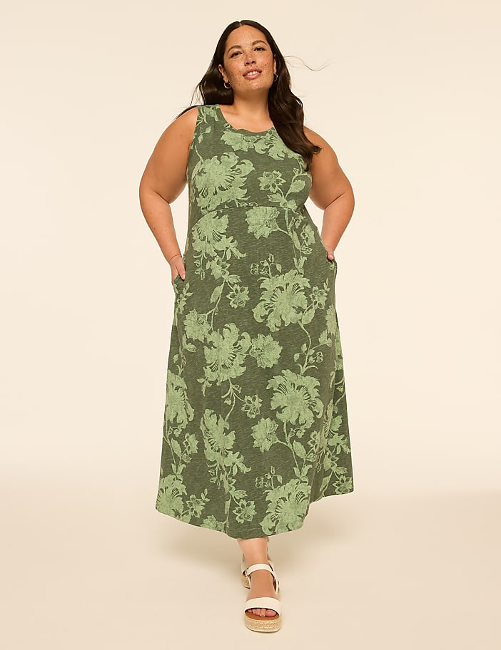 A woman in a floral green dress stands with her hands in her pockets against a plain, light-colored background.