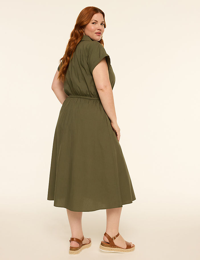 A woman in a green dress and sandals stands against a plain, light-colored background.