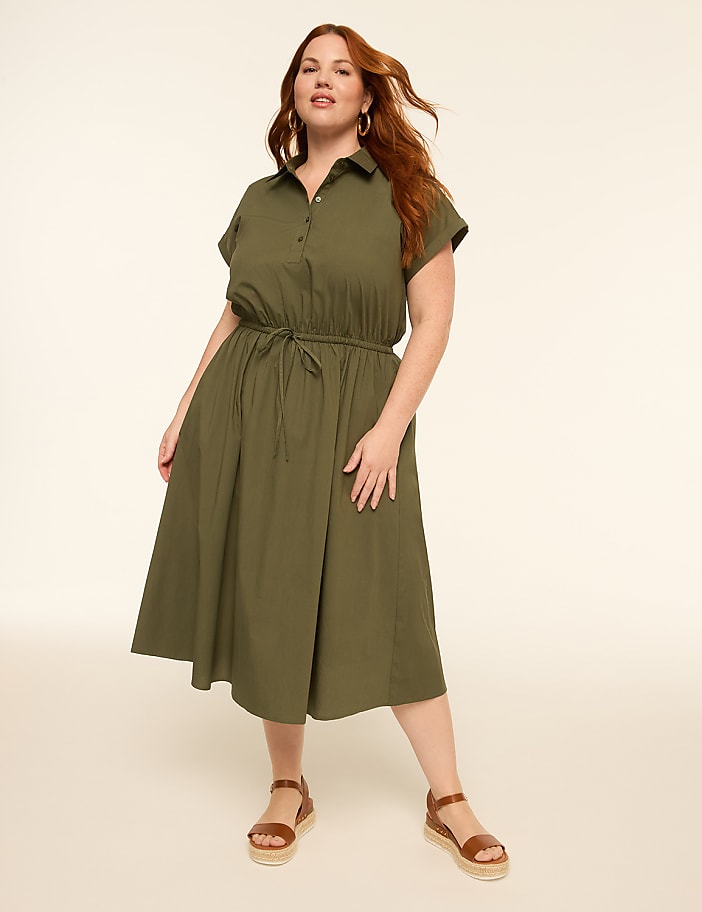 A woman in a green dress and sandals stands against a plain, light background.