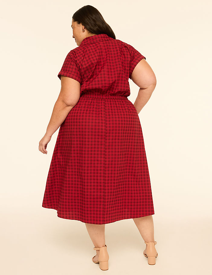 A woman in a red gingham dress stands against a plain, light-colored background.
