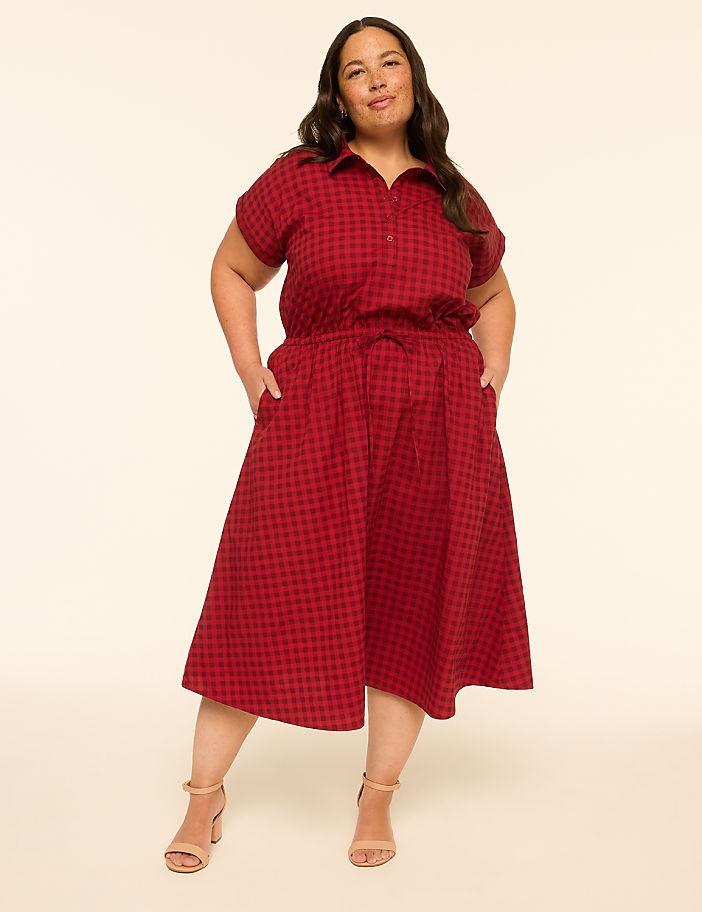 A woman in a red gingham dress stands with her hands in her pockets against a plain, light-colored background.
