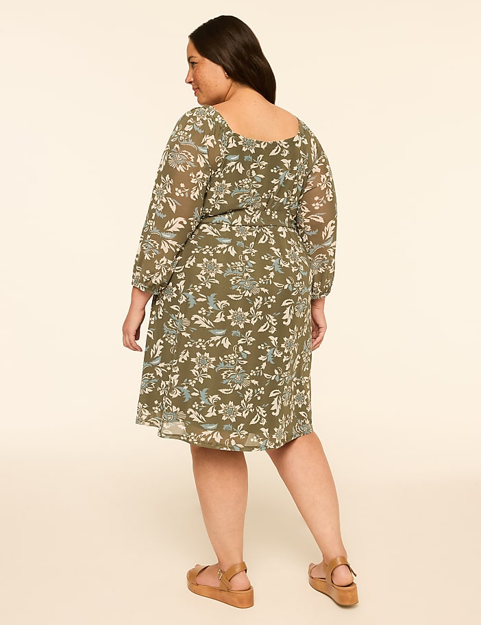 A woman in a floral dress stands against a plain, light-colored background.