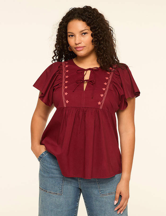 A woman in a maroon peasant blouse and blue jeans stands against a plain cream background.