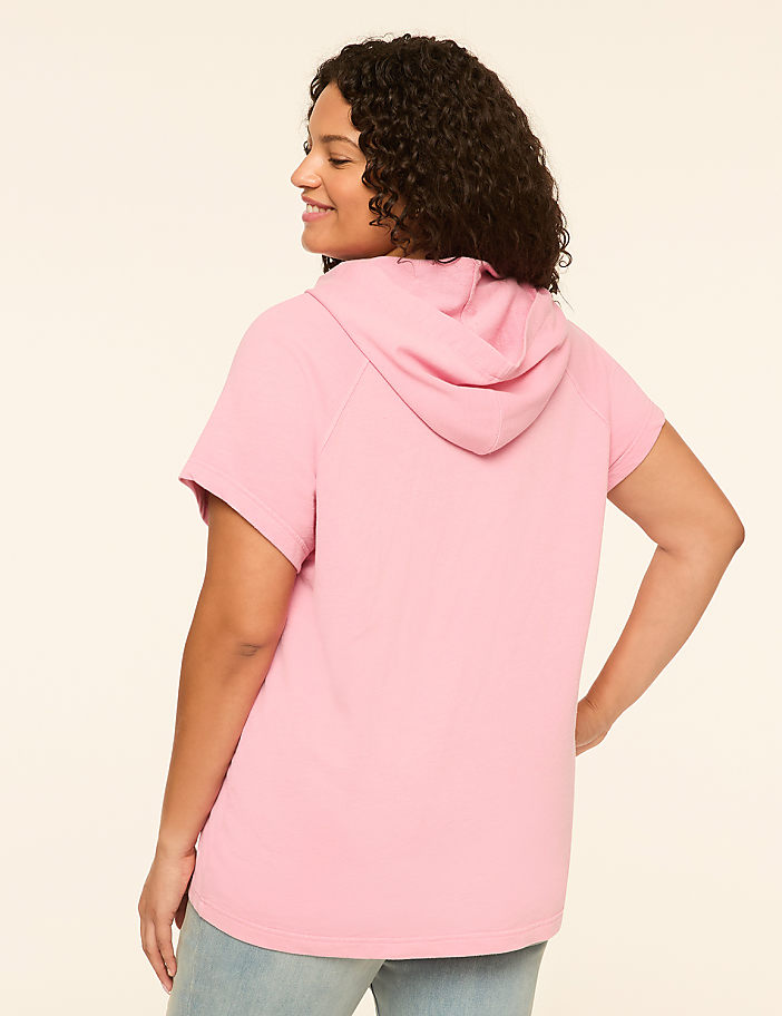 A woman in a pink short-sleeved hoodie stands against a plain, light-colored background.